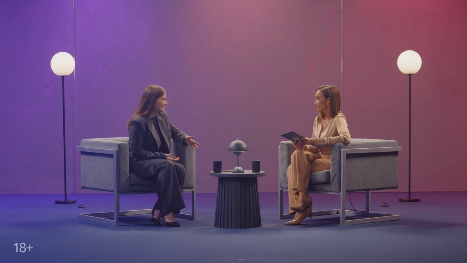 Still from the recording: two speakers in armchairs on a studio set lit purple and red