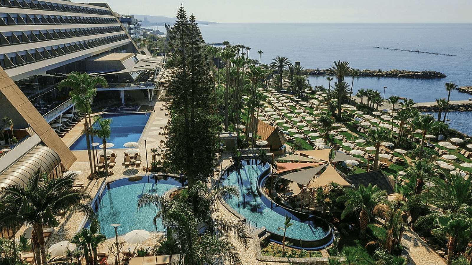 The beachfront hotel from above: the main building, pools among palm trees, a beach with umbrellas and the sea